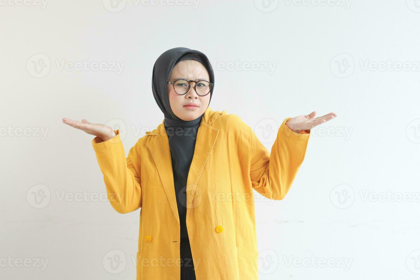 Beautiful young Asian Muslim woman in glasses, hijab and wearing yellow blazer with confused facial expression and both hands presenting empty space photo