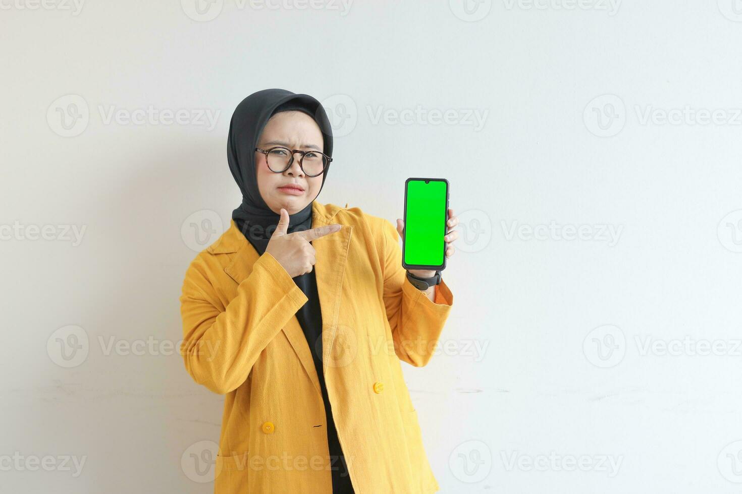 beautiful young asian woman in glasses, hijab and wearing yellow blazer is holding green screen mobile phone with sullen face photo
