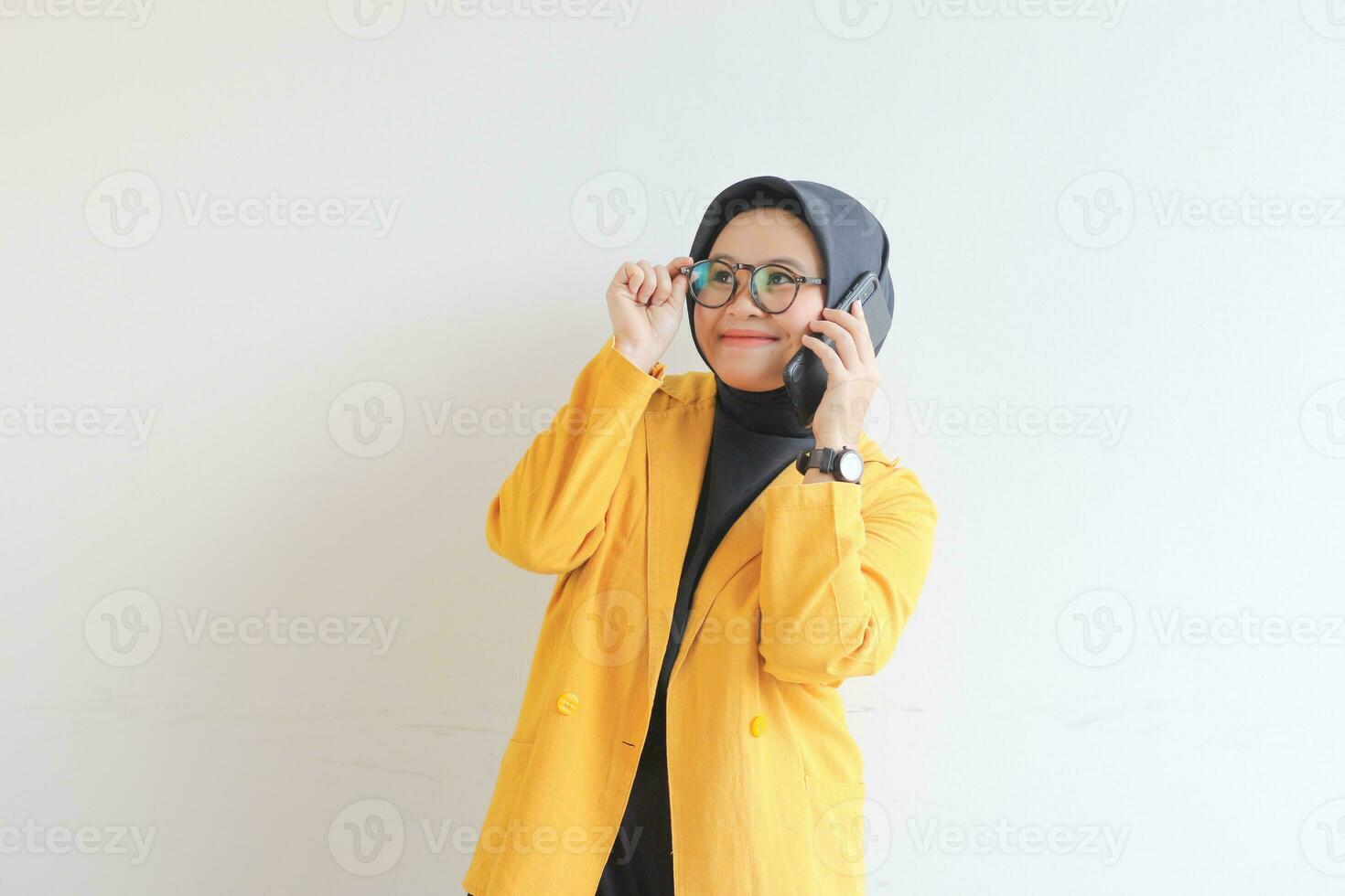 portrait of beautiful asian woman in hijab, glasses and wearing yellow blazer making phone call while smiling photo