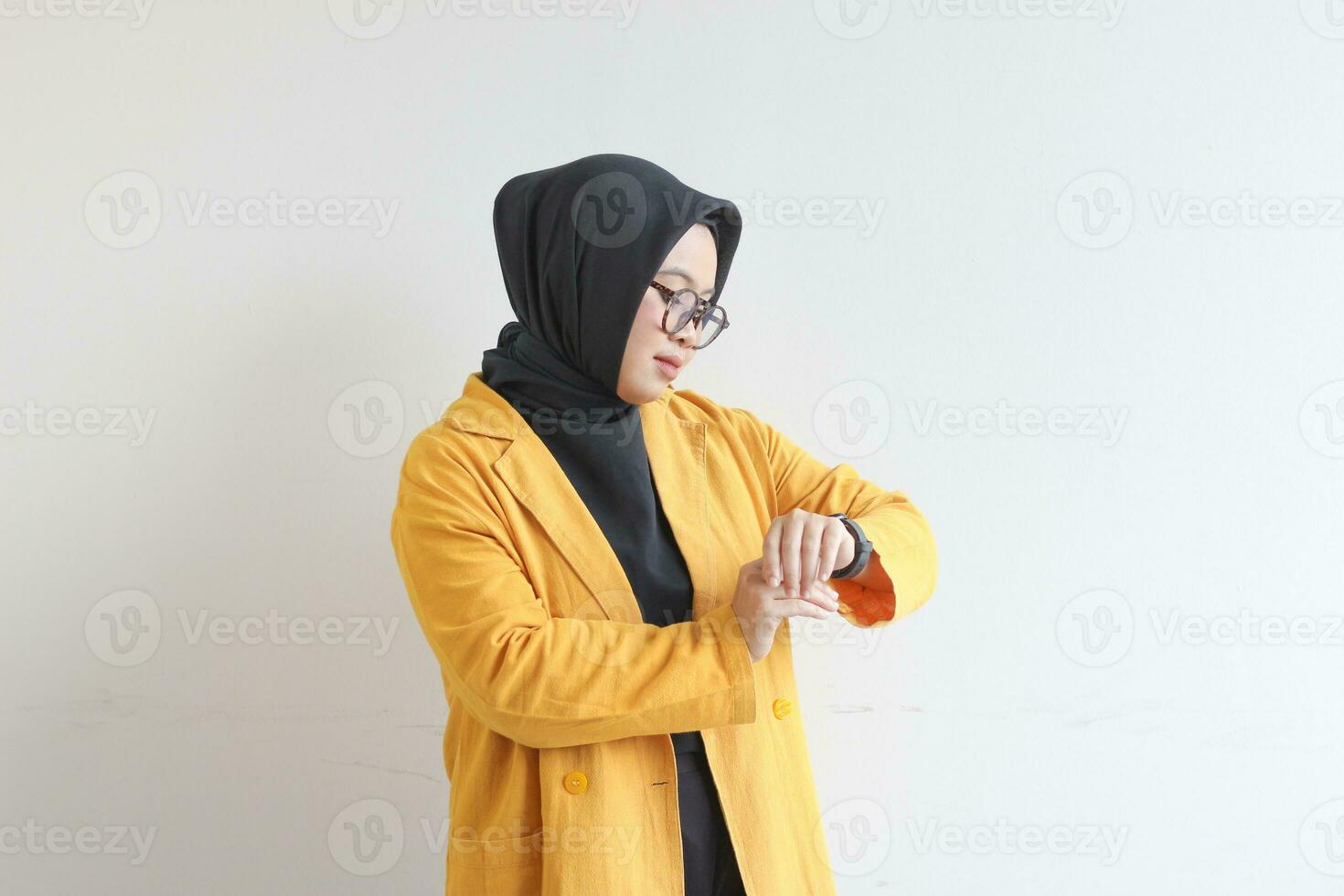 Beautiful young Asian Muslim woman, wearing glasses and yellow blazer gesturing at watch photo