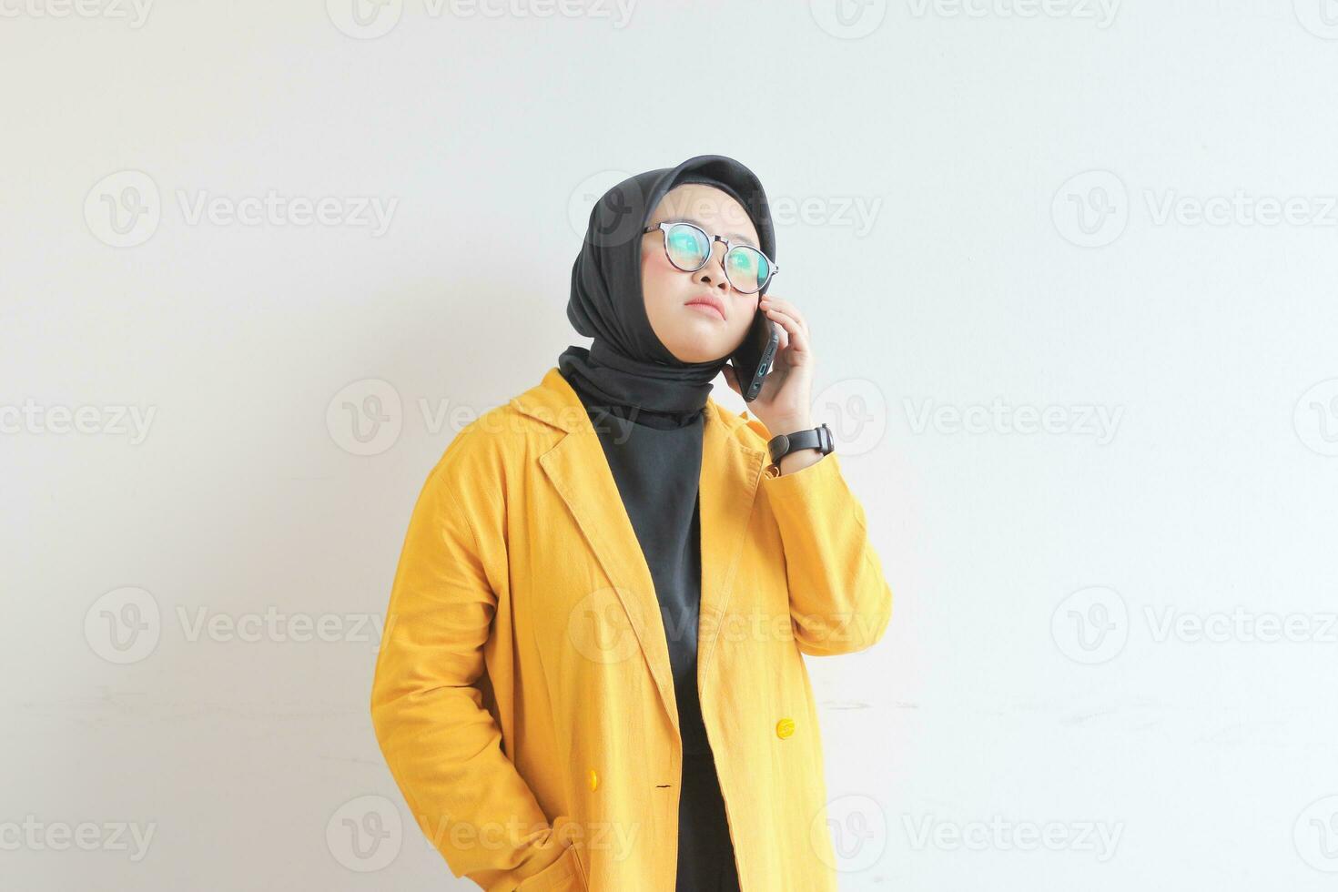portrait of beautiful asian woman in hijab, glasses and wearing yellow blazer making phone call while thinking photo
