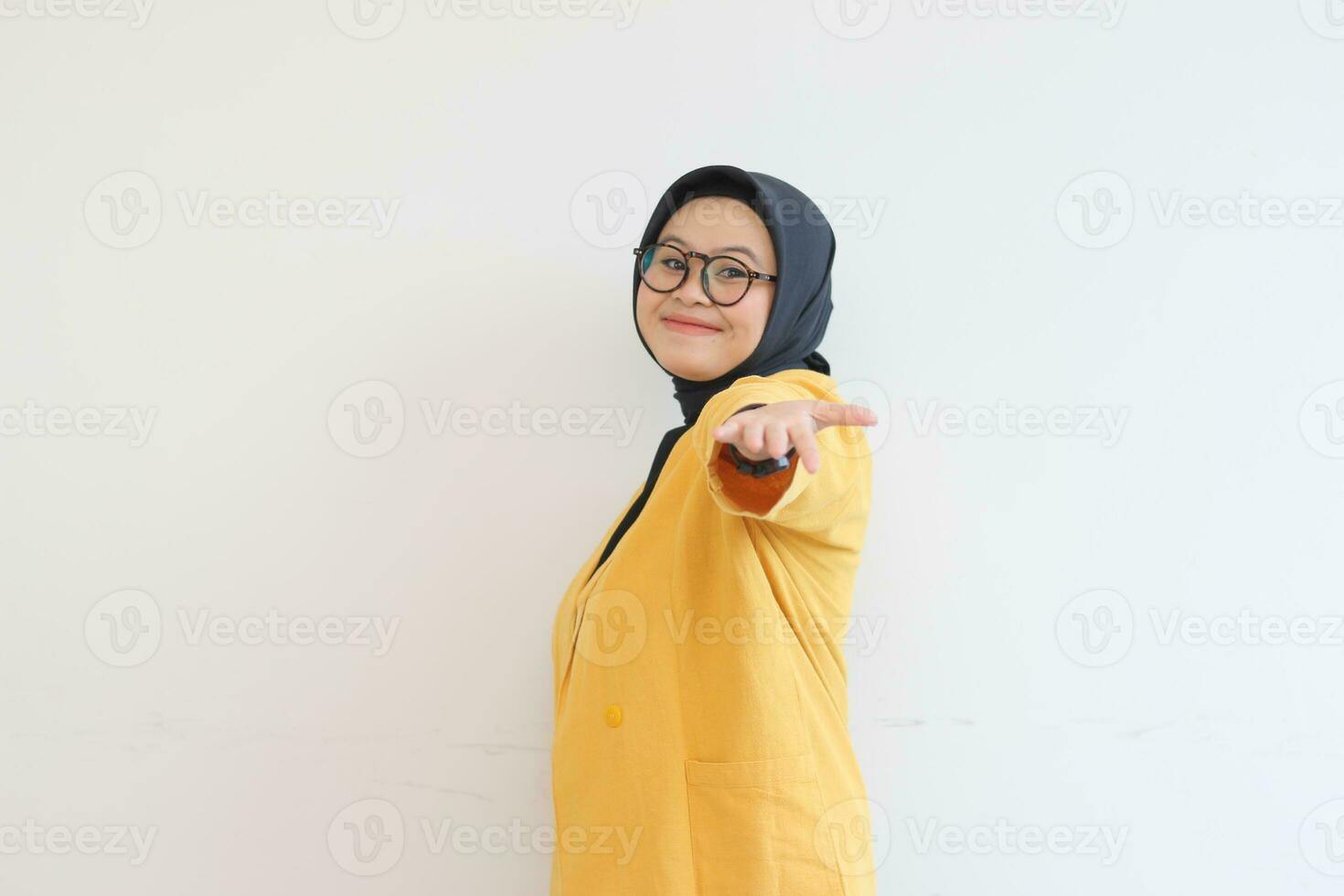 Beautiful young Asian Muslim woman in glasses, hijab and wearing yellow blazer smiling confident and happy with single hands presenting empty space photo