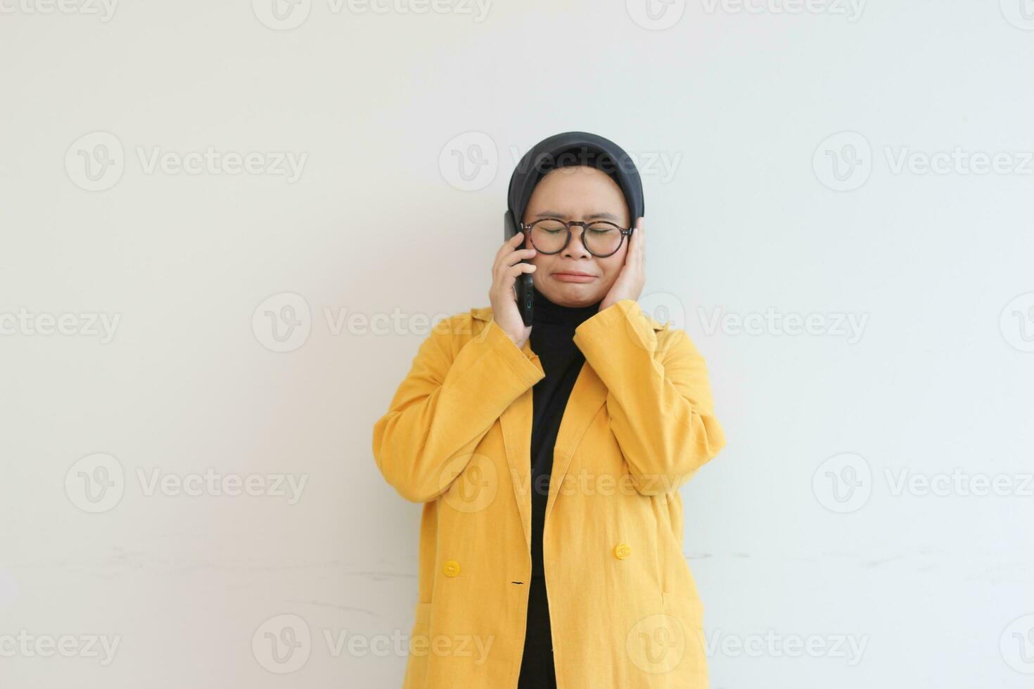 portrait of beautiful asian woman in hijab, glasses and wearing yellow blazer making phone call with sad face expression photo