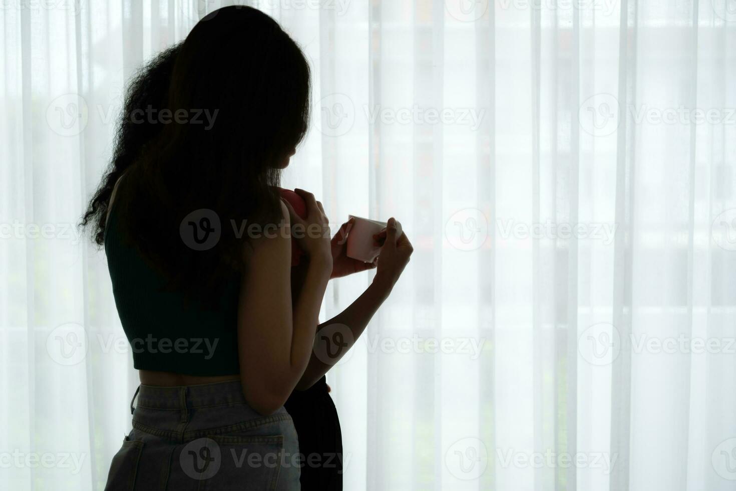 Both of cheerful young women drinking coffee at home holding coffee cups and chatting behind the white curtains of the living room where light shines photo