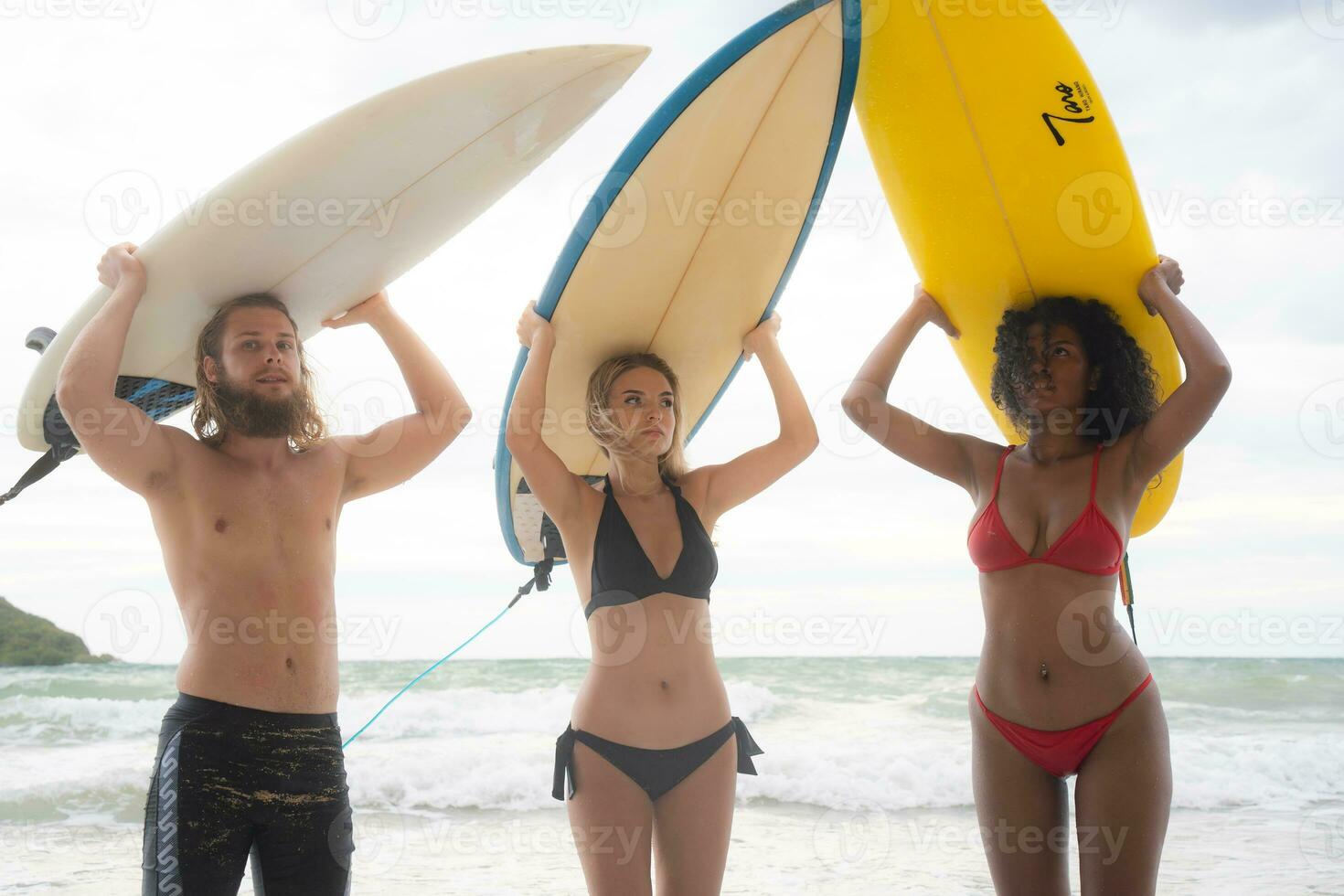 Rear view of two women and young man holding surfboards on their heads and walk into the sea to surf photo