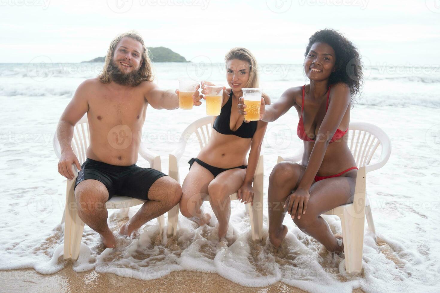 Multi-ethnic group of friends sitting on chairs with drinks on the beach photo