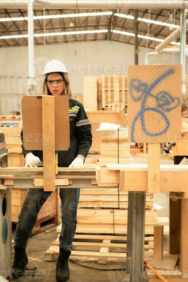 Female carpenter working in a woodworking factory, She is wearing a safety helmet and glasses. Checking the neatness of the planks against the design documents. photo