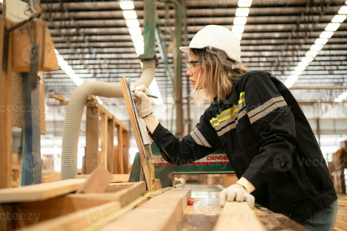 Female carpenter working in a woodworking factory, She is wearing a safety helmet and glasses. Checking the neatness of the planks against the design documents. photo