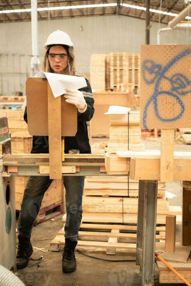 Female carpenter working in a woodworking factory, She is wearing a safety helmet and glasses. Checking the neatness of the planks against the design documents. photo