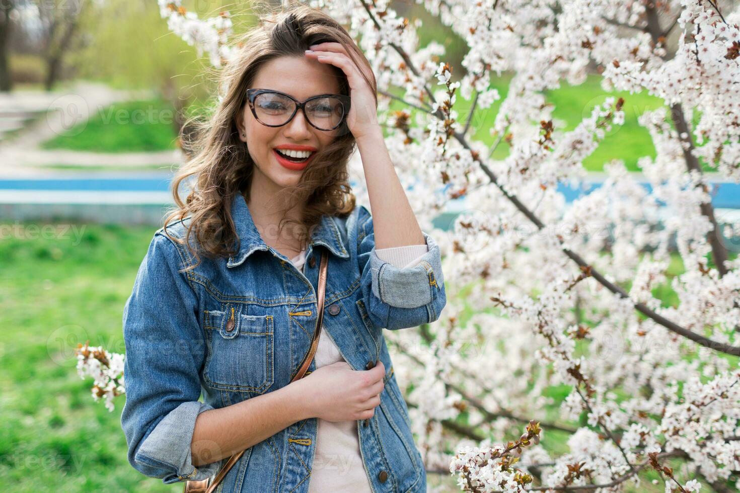 Sexy woman in jeans jacket, red full lips, wavy hairstyle posing near flower tree in spring day. photo