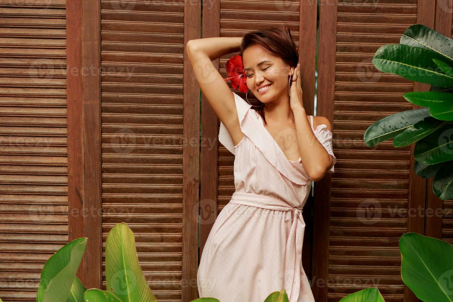 Graceful Asian woman with perfect skin  and hibiscus flower in hairs posing over wood wall and tropical plants. Summer fashion portrait. photo