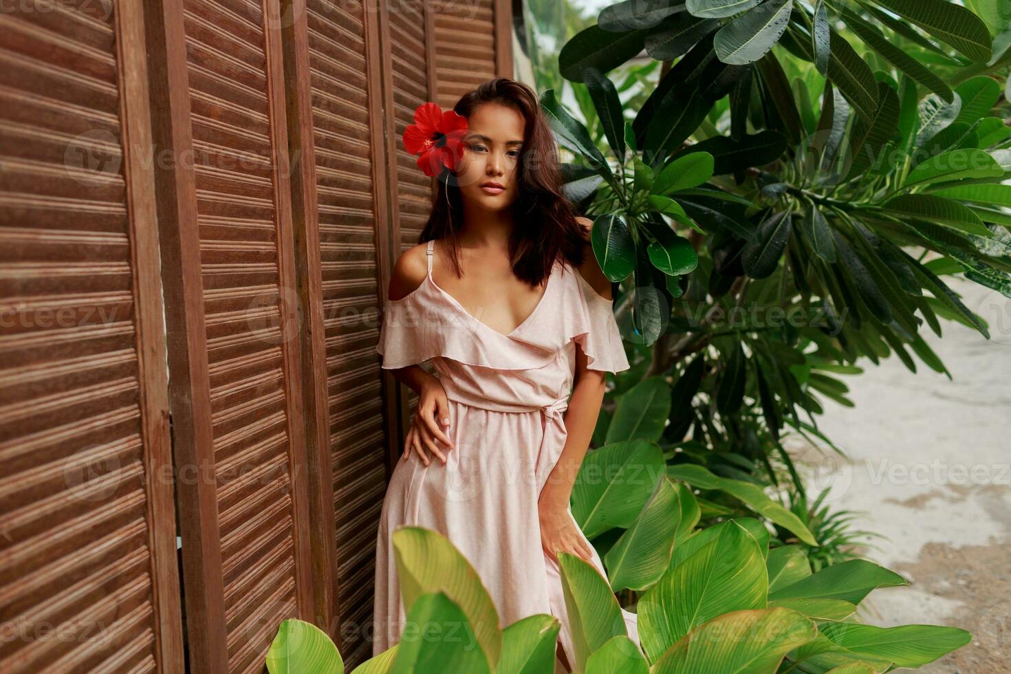 Graceful Asian woman with perfect skin  and hibiscus flower in hairs posing over wood wall and tropical plants. Summer fashion portrait. photo