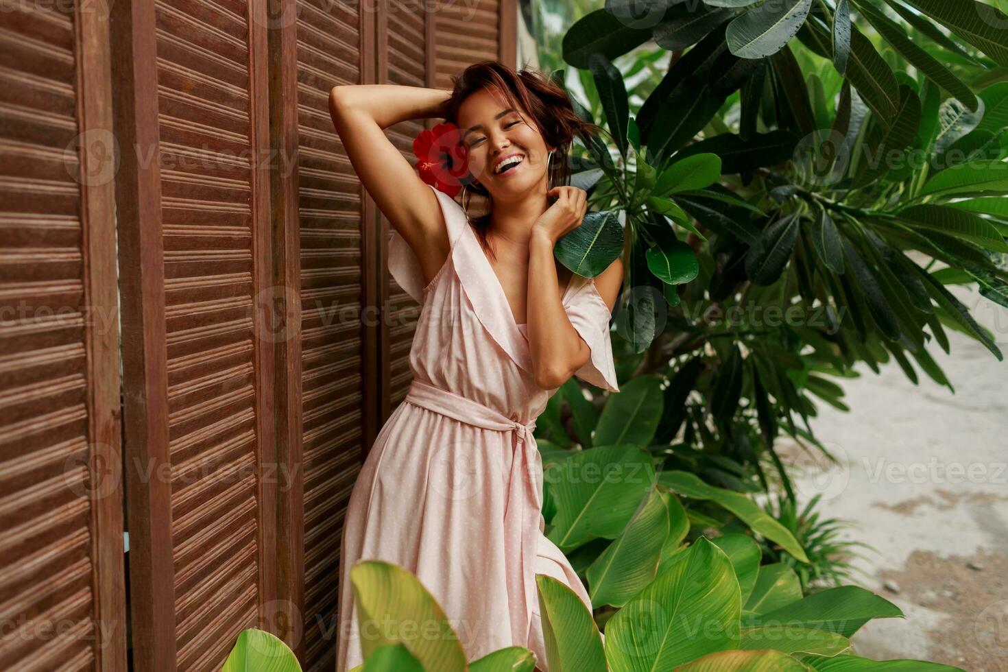 Graceful Asian woman with perfect skin  and hibiscus flower in hairs posing over wood wall and tropical plants. Summer fashion portrait. photo