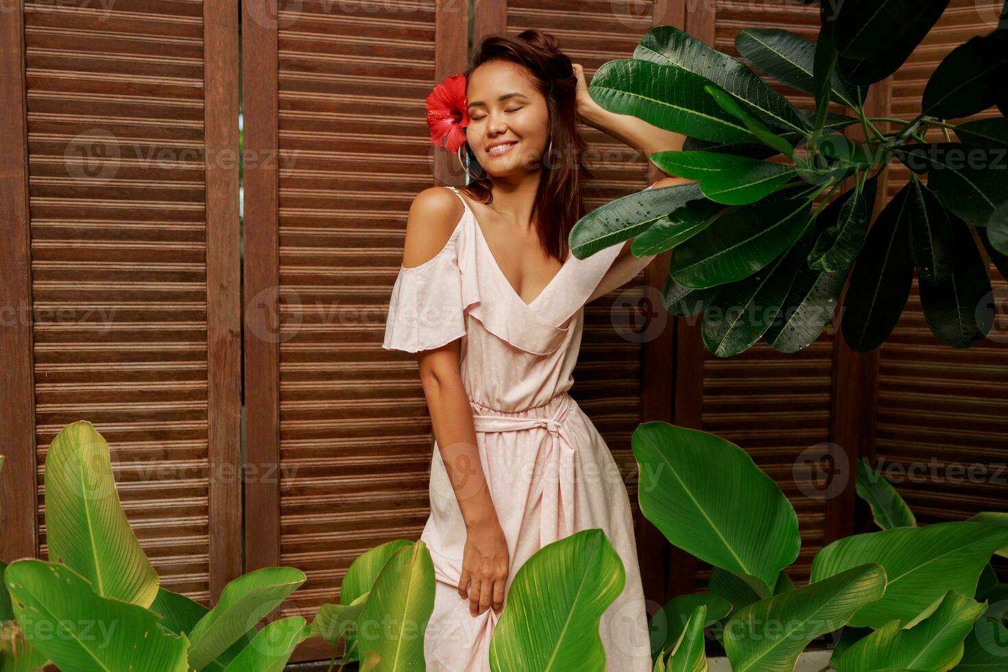 Graceful Asian woman with perfect skin  and hibiscus flower in hairs posing over wood wall and tropical plants. Summer fashion portrait. photo
