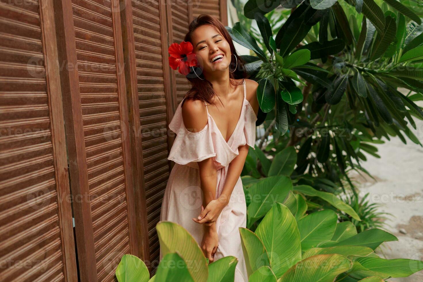 Graceful Asian woman with perfect skin  and hibiscus flower in hairs posing over wood wall and tropical plants. Summer fashion portrait. photo