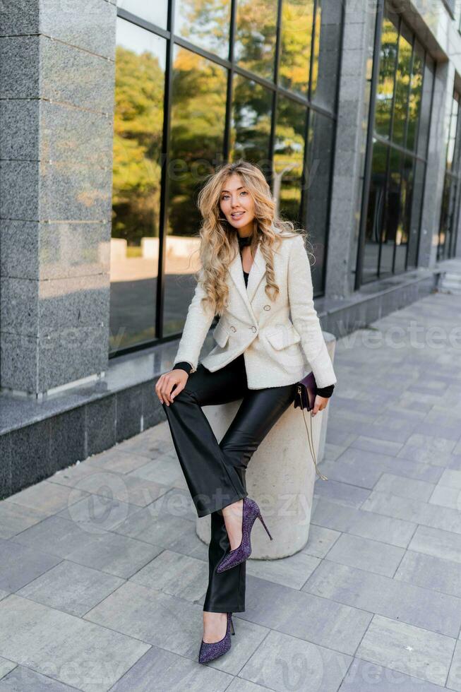 Outdoor photo of sucsessful blond woman with perfect wavy hairs in casual  outfit with luxury purse posing over modern business center.