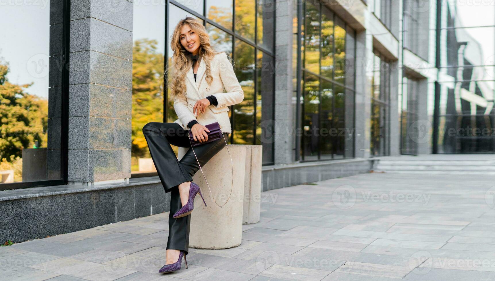Outdoor photo of sucsessful blond woman with perfect wavy hairs in casual  outfit with luxury purse posing over modern business center.