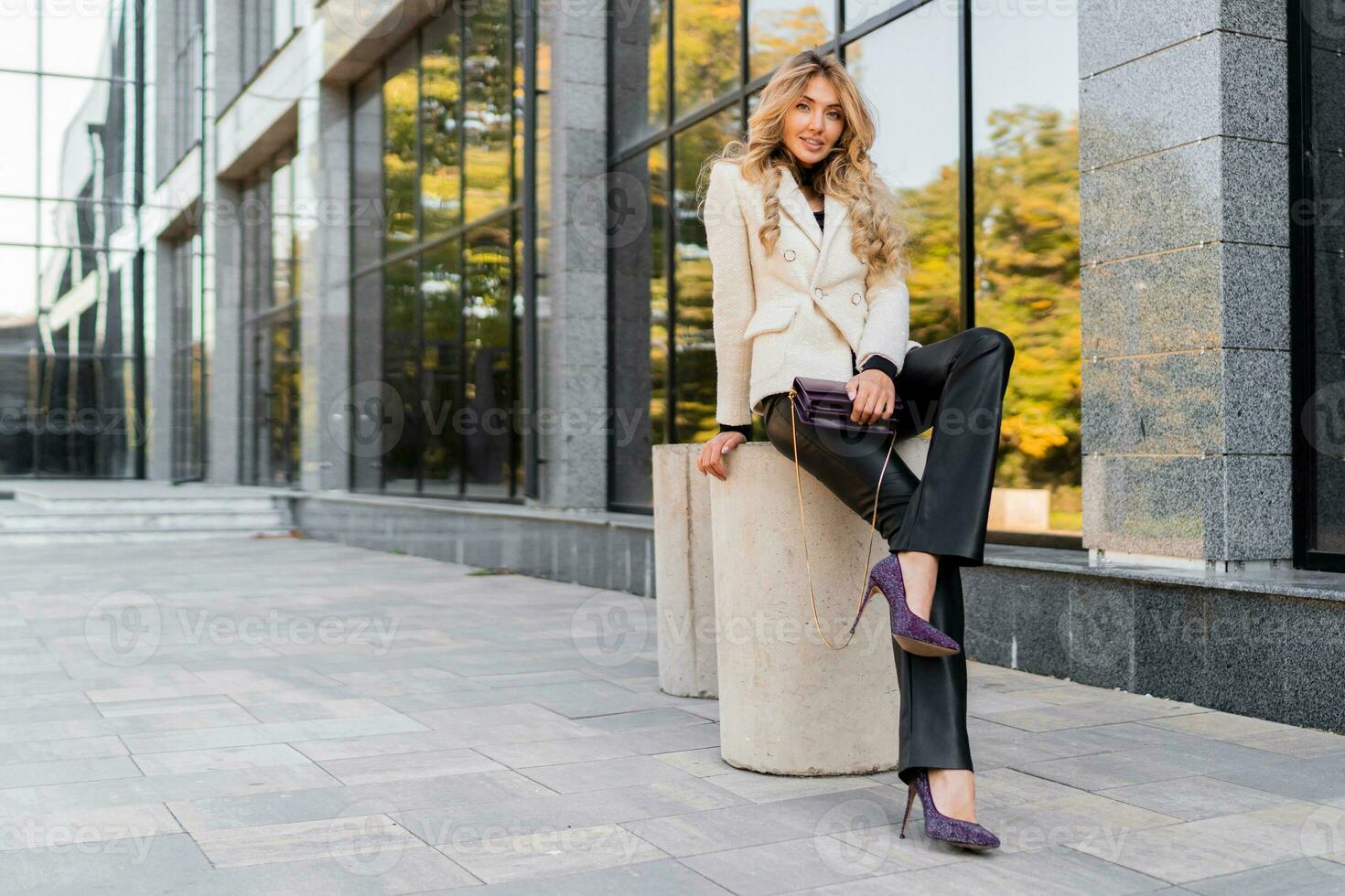 Outdoor photo of sucsessful blond woman with perfect wavy hairs in casual  outfit with luxury purse posing over modern business center.