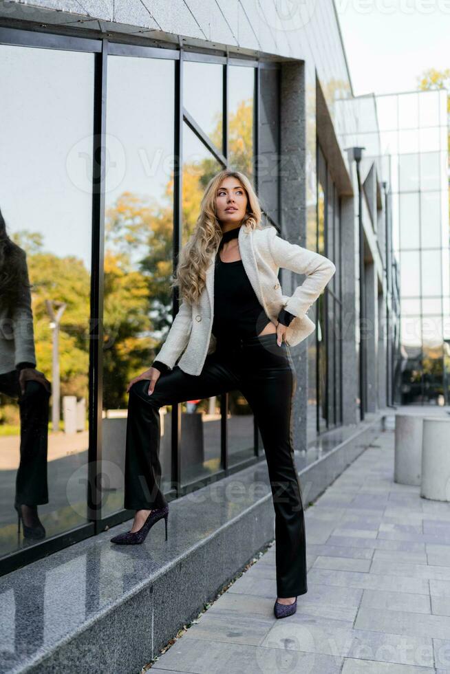 elegante bonito rubio mujer con Perfecto ondulado pelos y lleno labios en casual chaqueta posando terminado moderno calle. foto