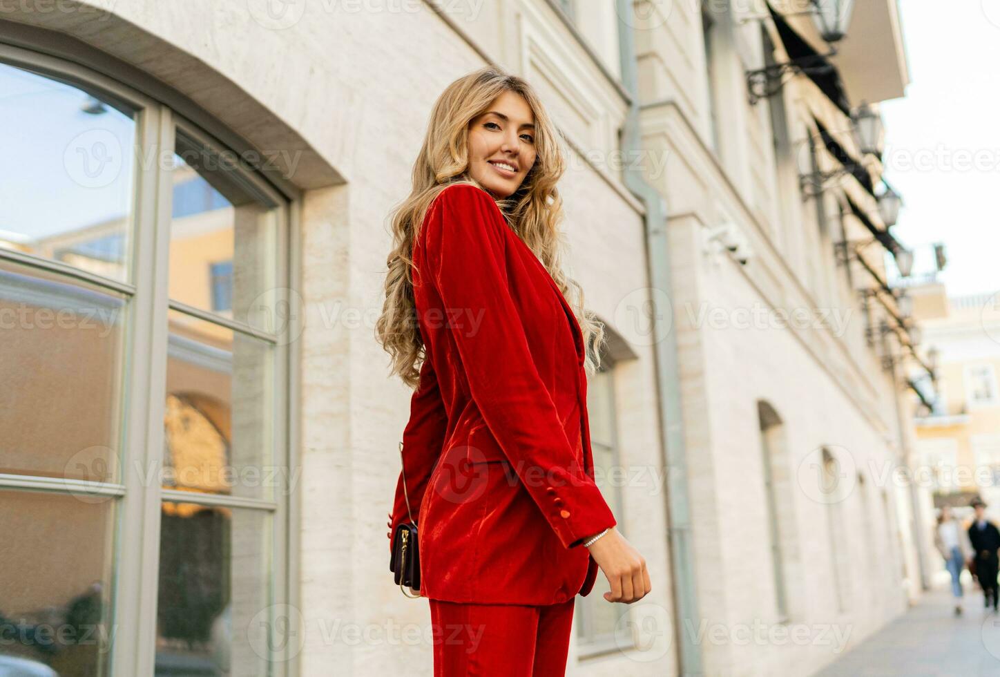 Beautiful smiling woman  in  elegant red velvet suit holding purse and  posin outdoor in old european city.    Blond wavy hairs, perfet skin, full lips. photo