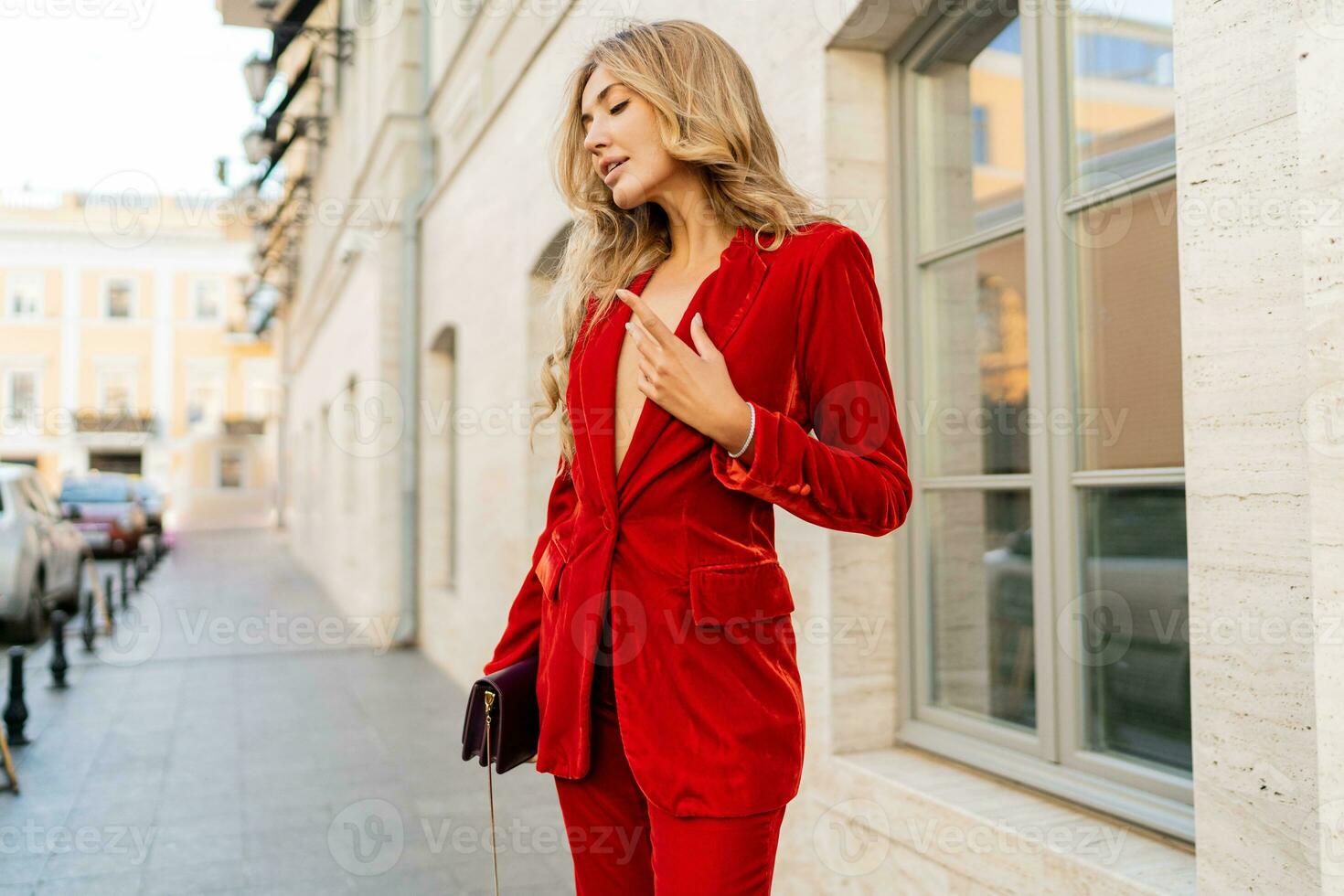 Beautiful smiling woman  in  elegant red velvet suit holding purse and  posin outdoor in old european city.    Blond wavy hairs, perfet skin, full lips. photo