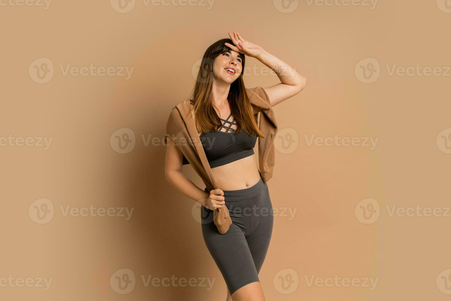 Brunette woman in stylish sport outfit   posing in studio on beige bakground. Grey crop top and leggins. photo