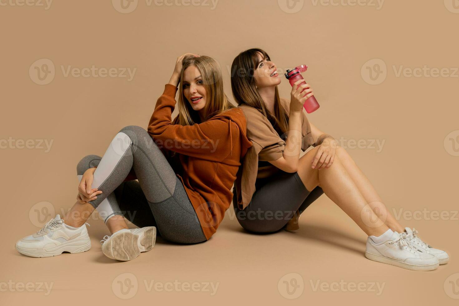 Two laughing sportive  women chilling after training,  sitting on floor in studio on beige background. photo