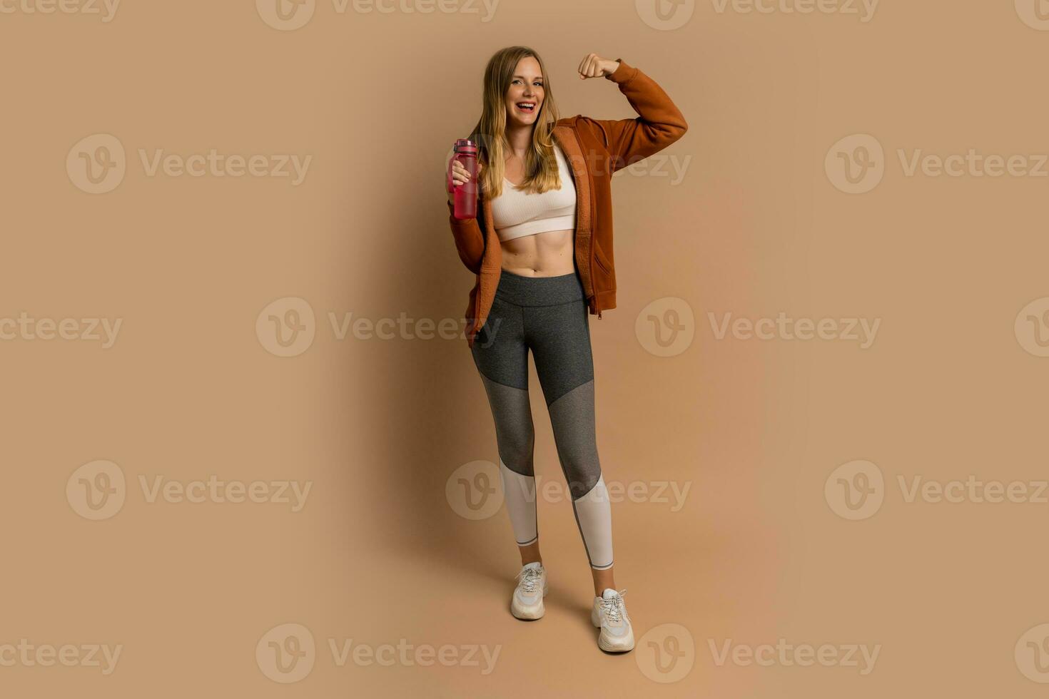 Pretty blond woman in sportive outfit  posing on beige background.  Holding bottle of water. Full lenght. photo