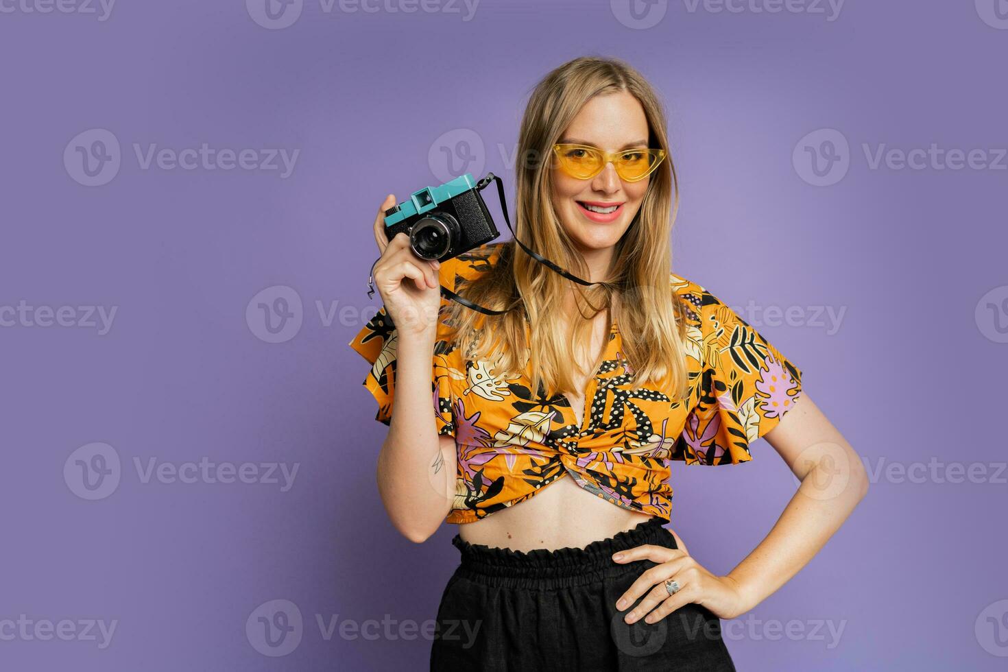 Pretty blond woman in stylish summer outfit holding camera and posing on purple background in studio. Travel and summer fashion concept. photo