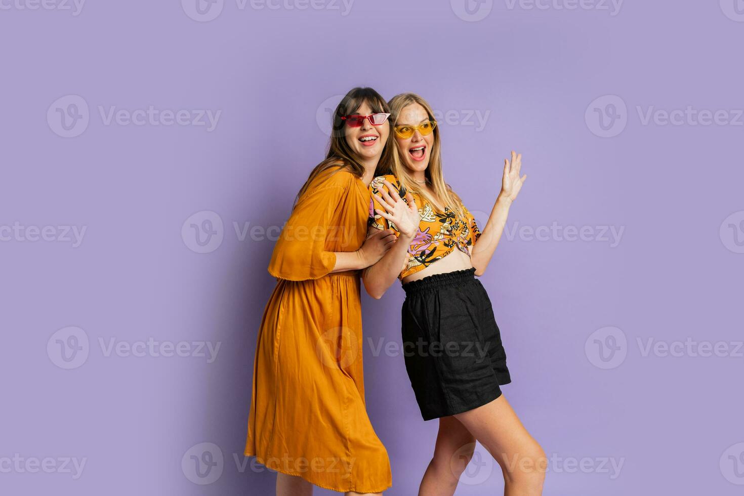 Two playful stylish women posing in studio over purple background. Friends dancing and having fun. photo