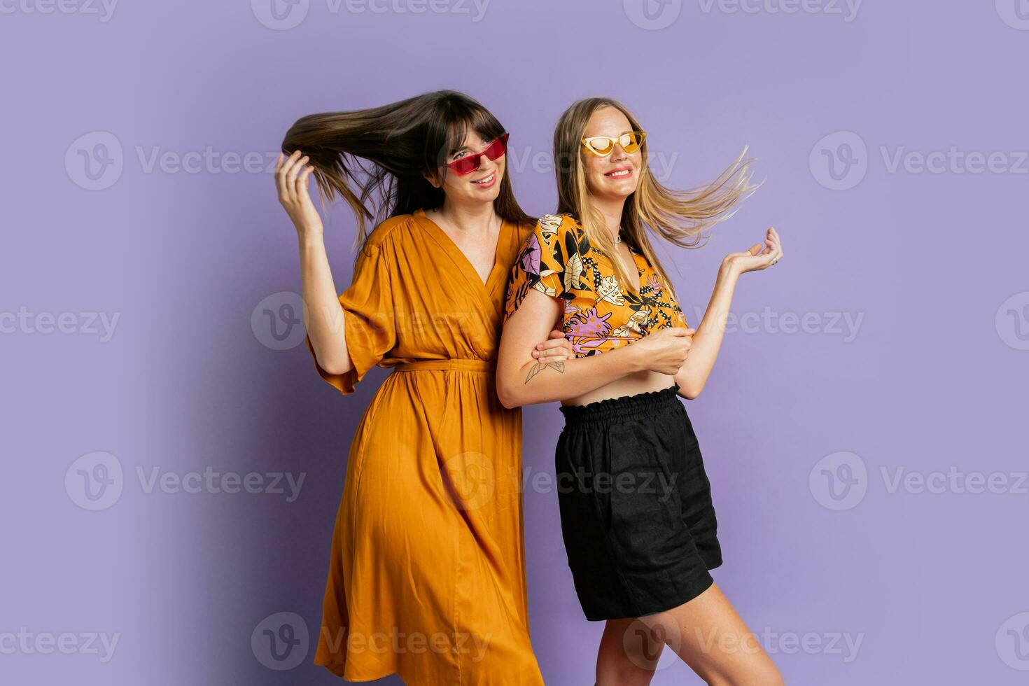Two playful stylish women posing in studio over purple background. Friends dancing and having fun. photo