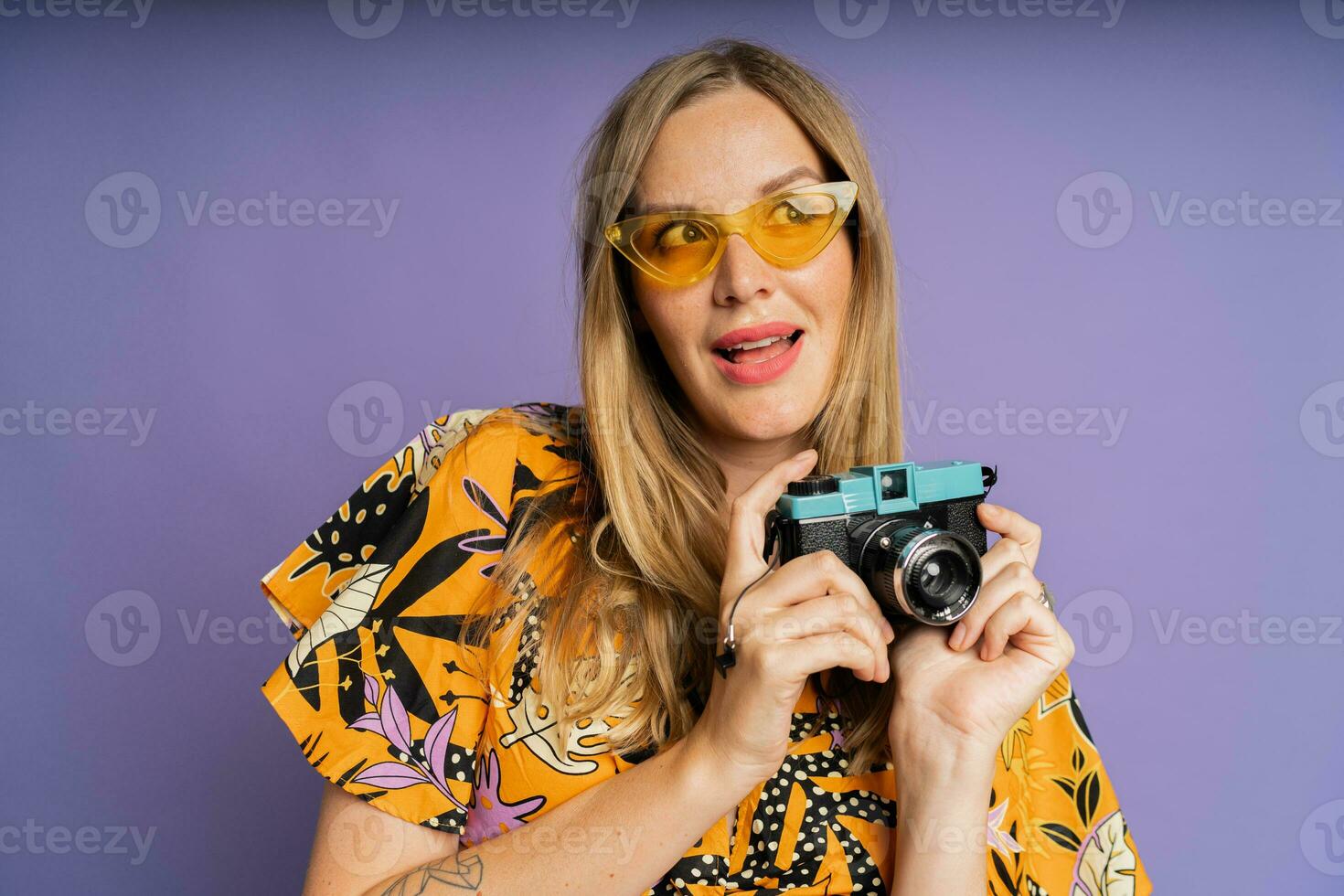 Pretty blond woman in stylish summer outfit holding camera and posing on purple background in studio. Travel and summer fashion concept. photo