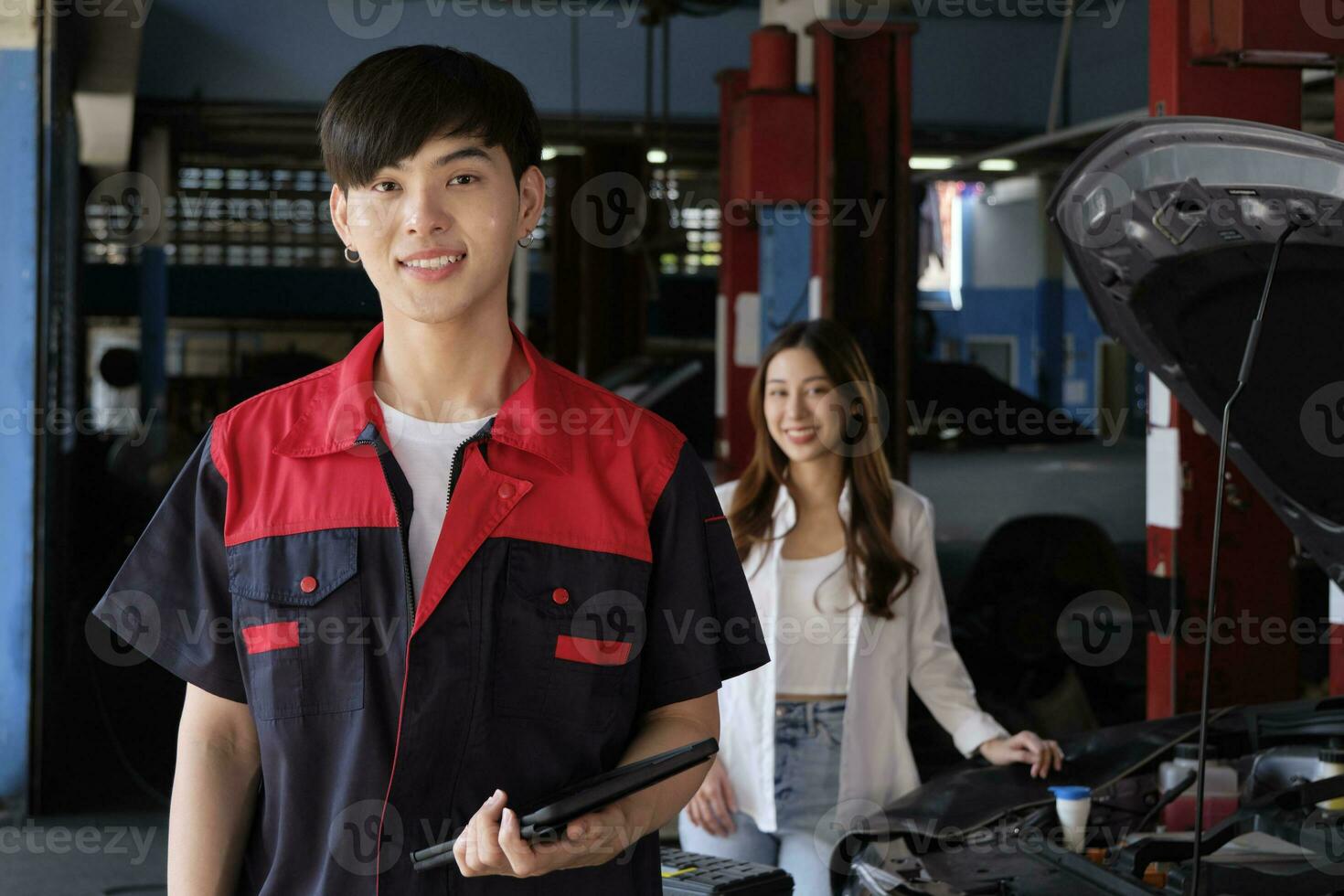 Portrait of young Asian male automotive mechanic worker with female customer's EV car at maintenance garage, happy smiles, professional service station, check and repair in auto transport industry. photo