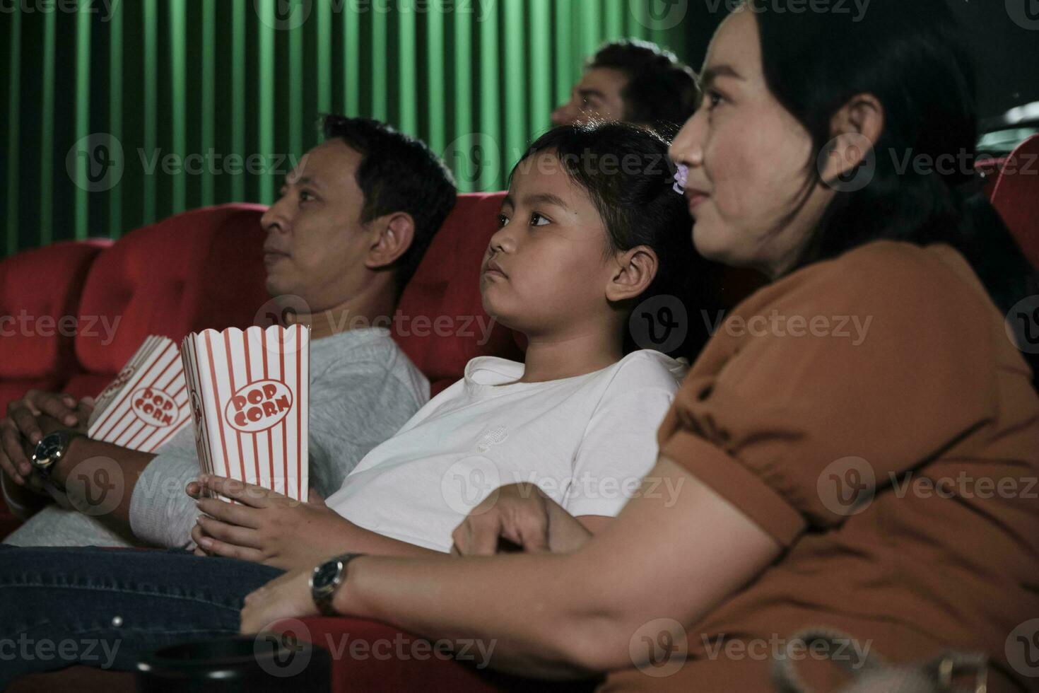 Asian family audience enjoys watching cinema together at movie theaters. Kid and parents have indoor entertainment lifestyle with performance art shows, happy and cheerful with popcorn and a smile. photo