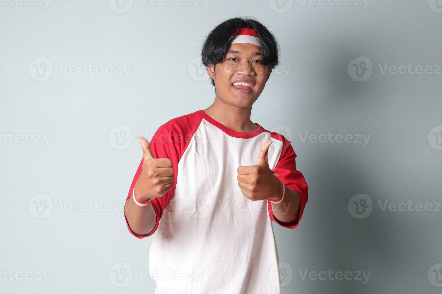 retrato de atractivo asiático hombre en camiseta con rojo y blanco cinta en cabeza, demostración bueno trabajo mano gesto con pulgares arriba. aislado imagen en gris antecedentes foto