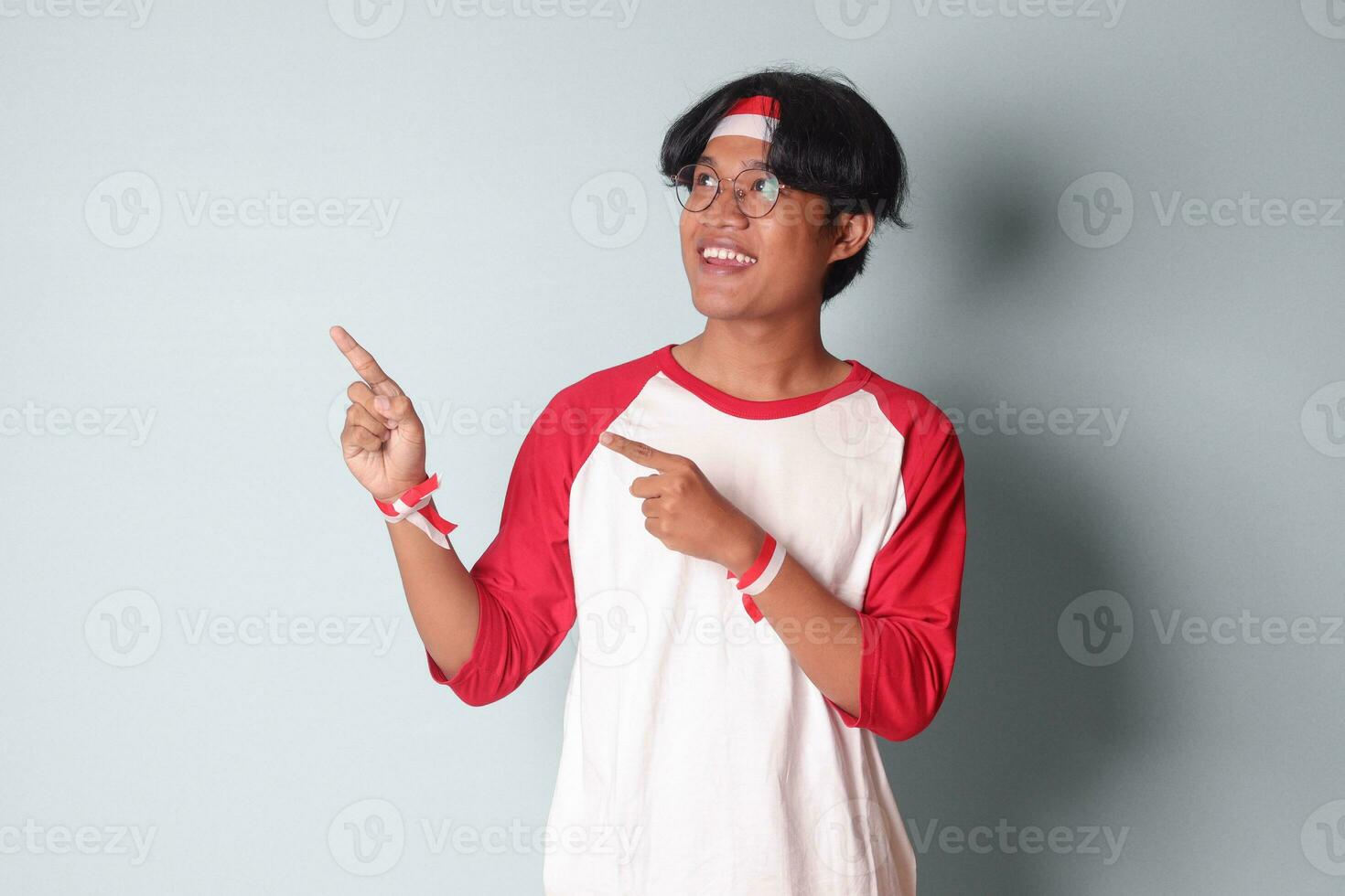 Portrait of attractive Asian man in t-shirt with red and white ribbon on head, pointing at something with finger. Isolated image on gray background photo