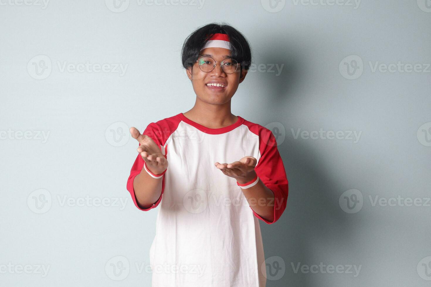 Portrait of attractive Asian man in t-shirt with red and white ribbon on head, pulling hands towards camera, inviting someone to come inside, welcoming gesture. Isolated image on gray background photo