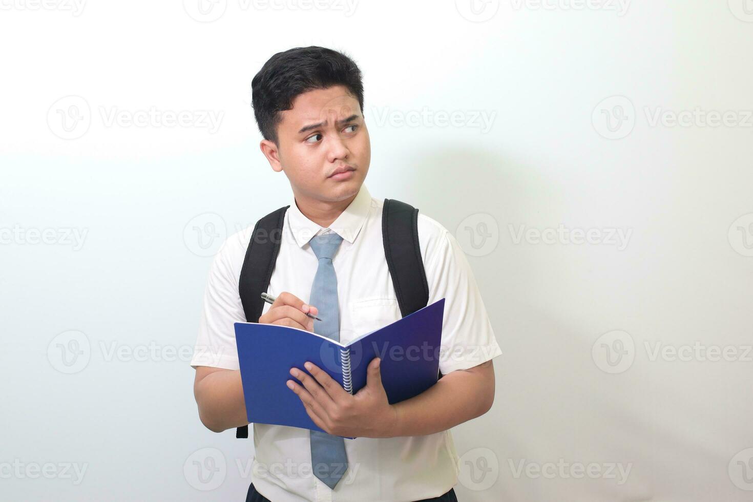 indonesio mayor alto colegio estudiante vistiendo blanco camisa uniforme con gris Corbata escritura en Nota libro utilizando bolígrafo y pensando acerca de un idea. aislado imagen en blanco antecedentes foto