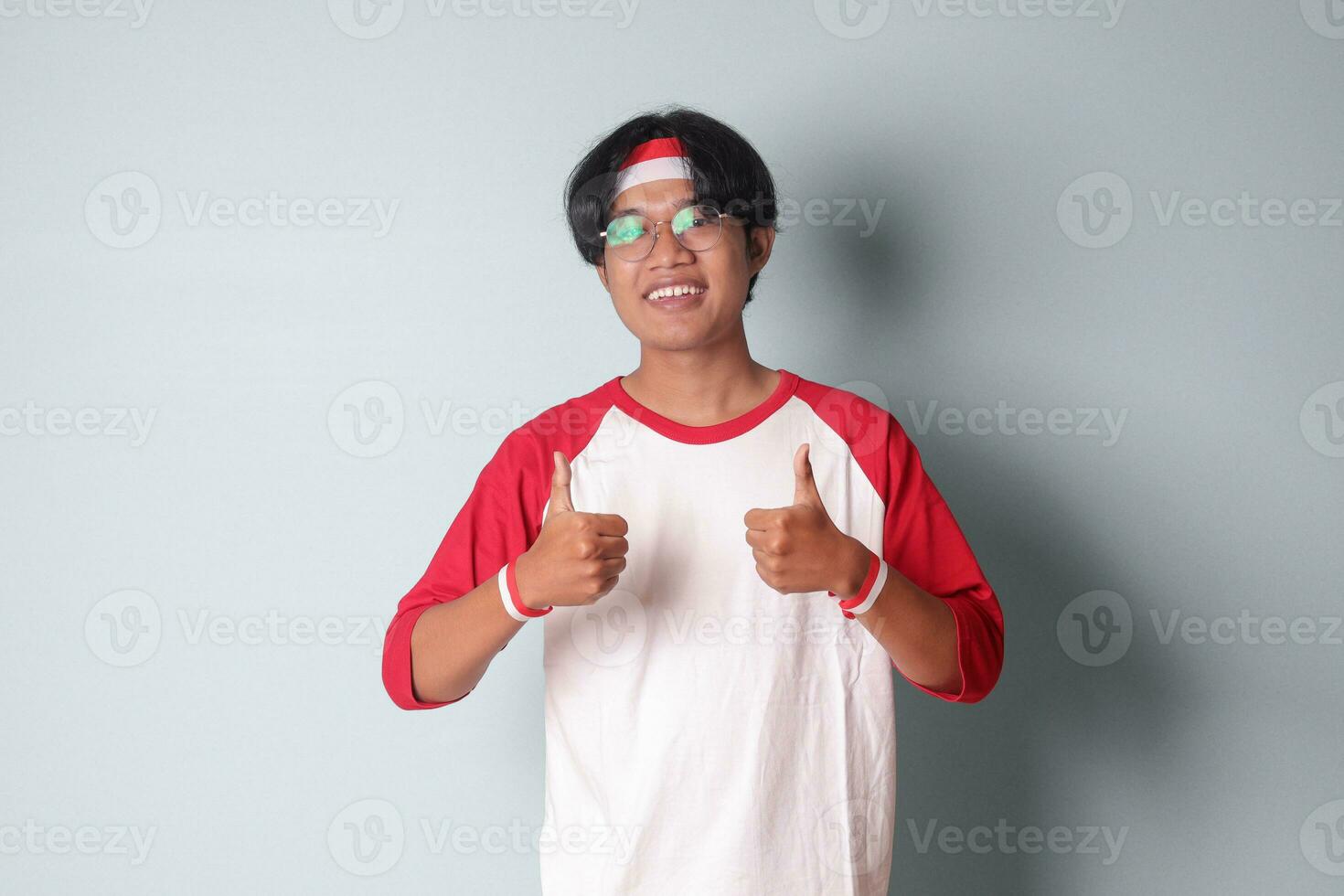 retrato de atractivo asiático hombre en camiseta con rojo y blanco cinta en cabeza, demostración bueno trabajo mano gesto con pulgares arriba. aislado imagen en gris antecedentes foto