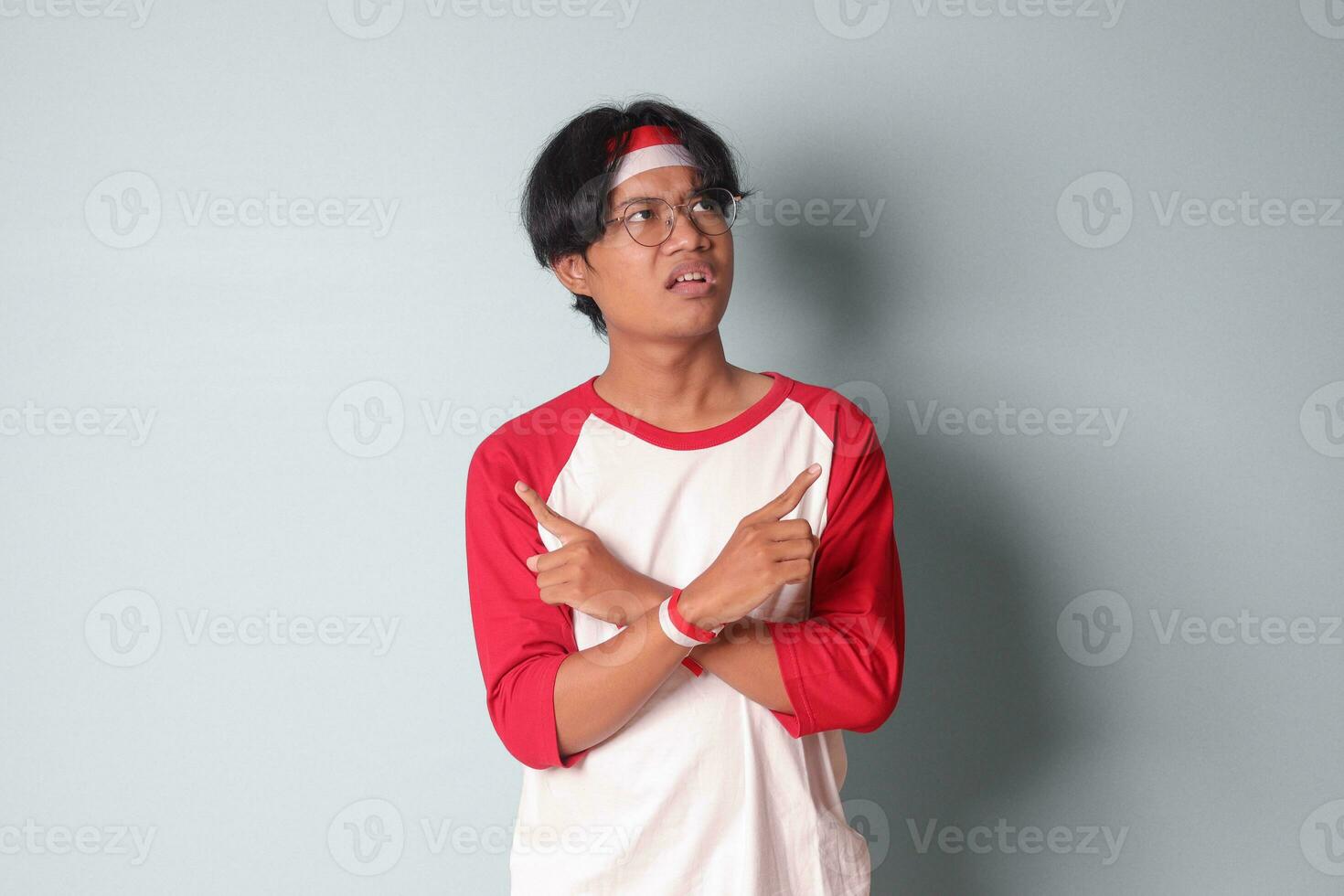 Portrait of attractive Asian man in t-shirt with red and white ribbon on head, choosing between 2 different options, holding two hands with empty space. Decision concept. Isolated image on gray photo
