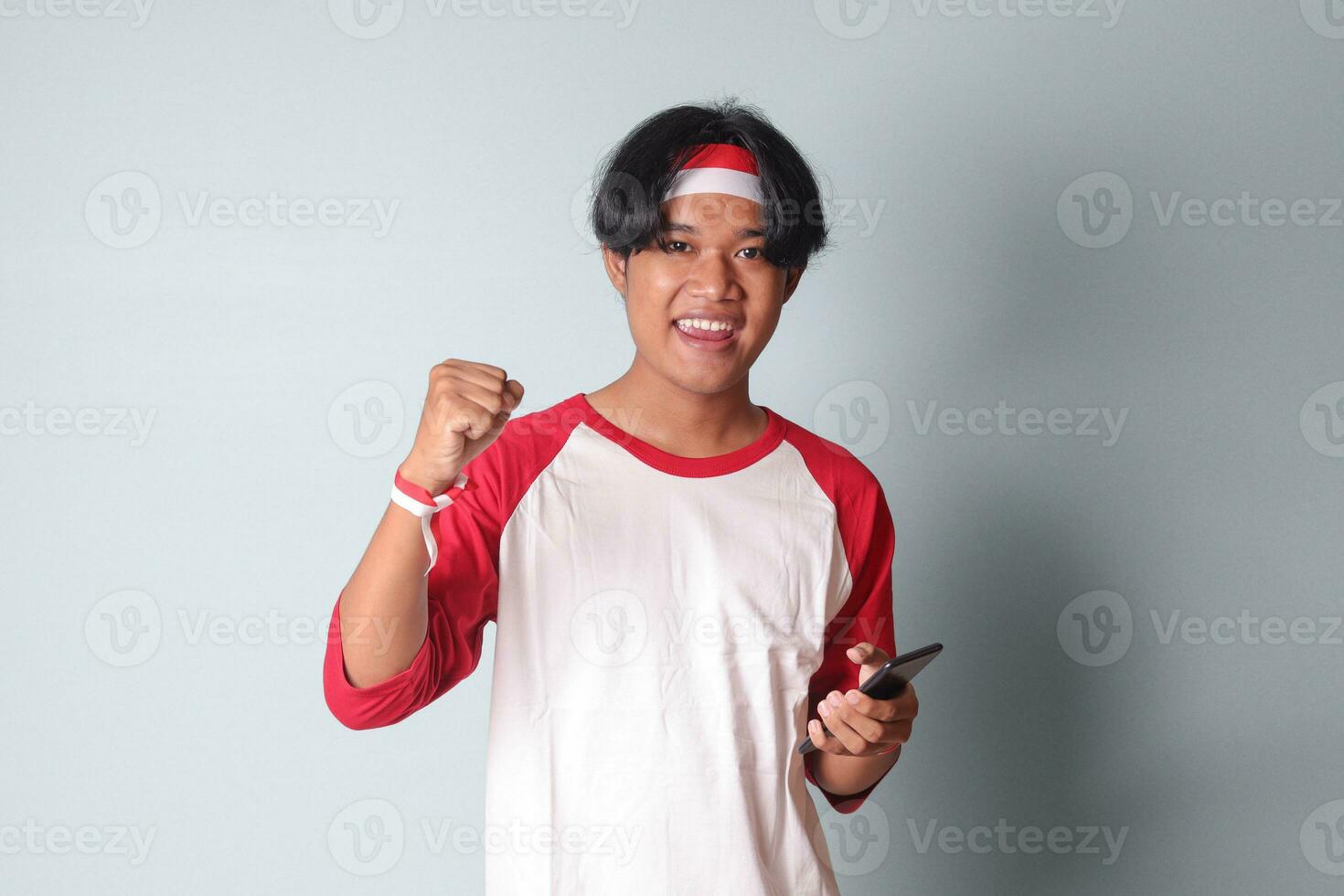 retrato de atractivo asiático hombre en camiseta con rojo y blanco cinta en cabeza, participación móvil teléfono mientras levantamiento su puño, celebrando éxito. aislado imagen en gris antecedentes foto