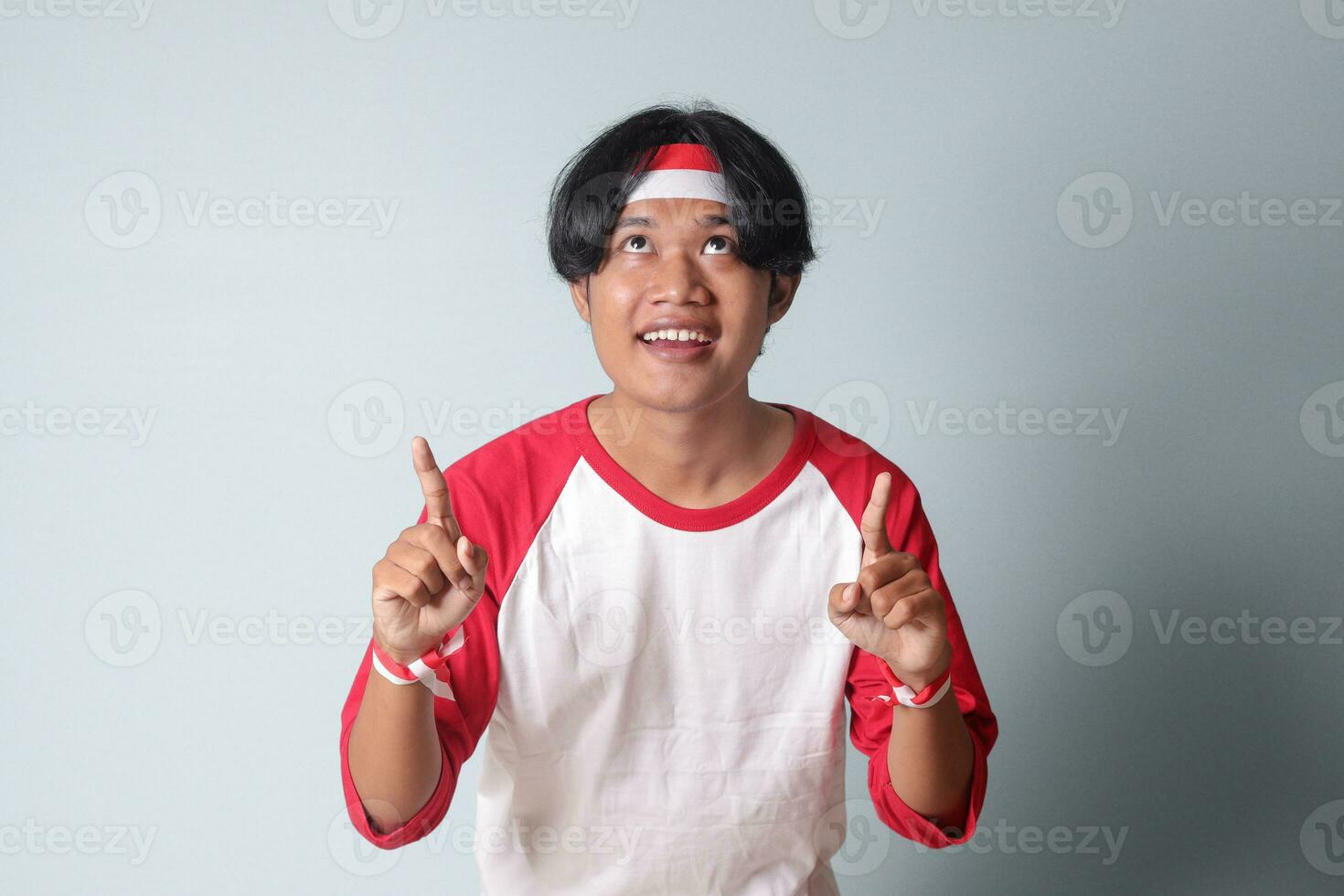 Portrait of attractive Asian man in t-shirt with red and white ribbon on head, pointing at something with finger. Isolated image on gray background photo