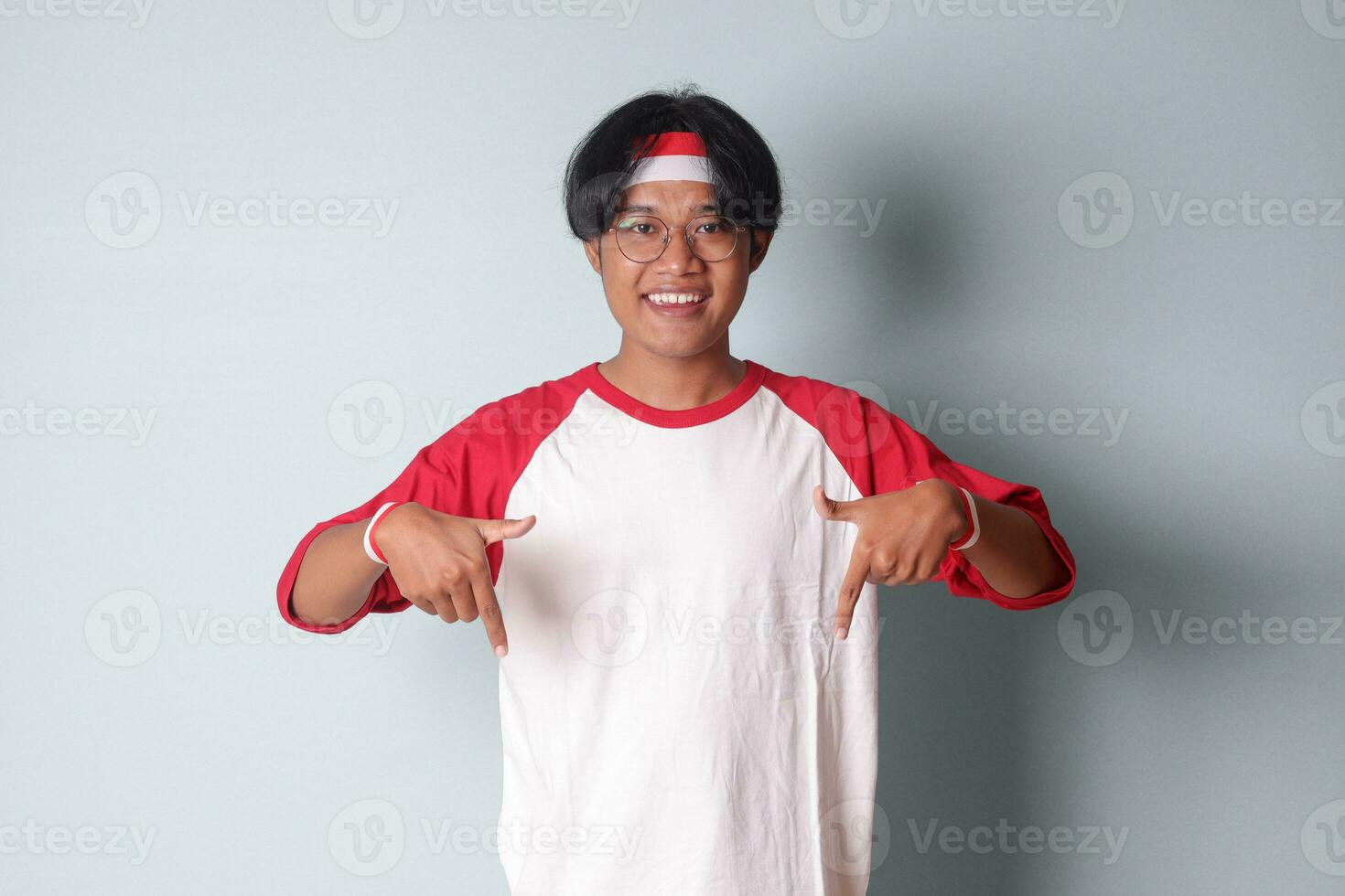 retrato de atractivo asiático hombre en camiseta con rojo y blanco cinta en cabeza, señalando a alguna cosa abajo con dedo. aislado imagen en gris antecedentes foto