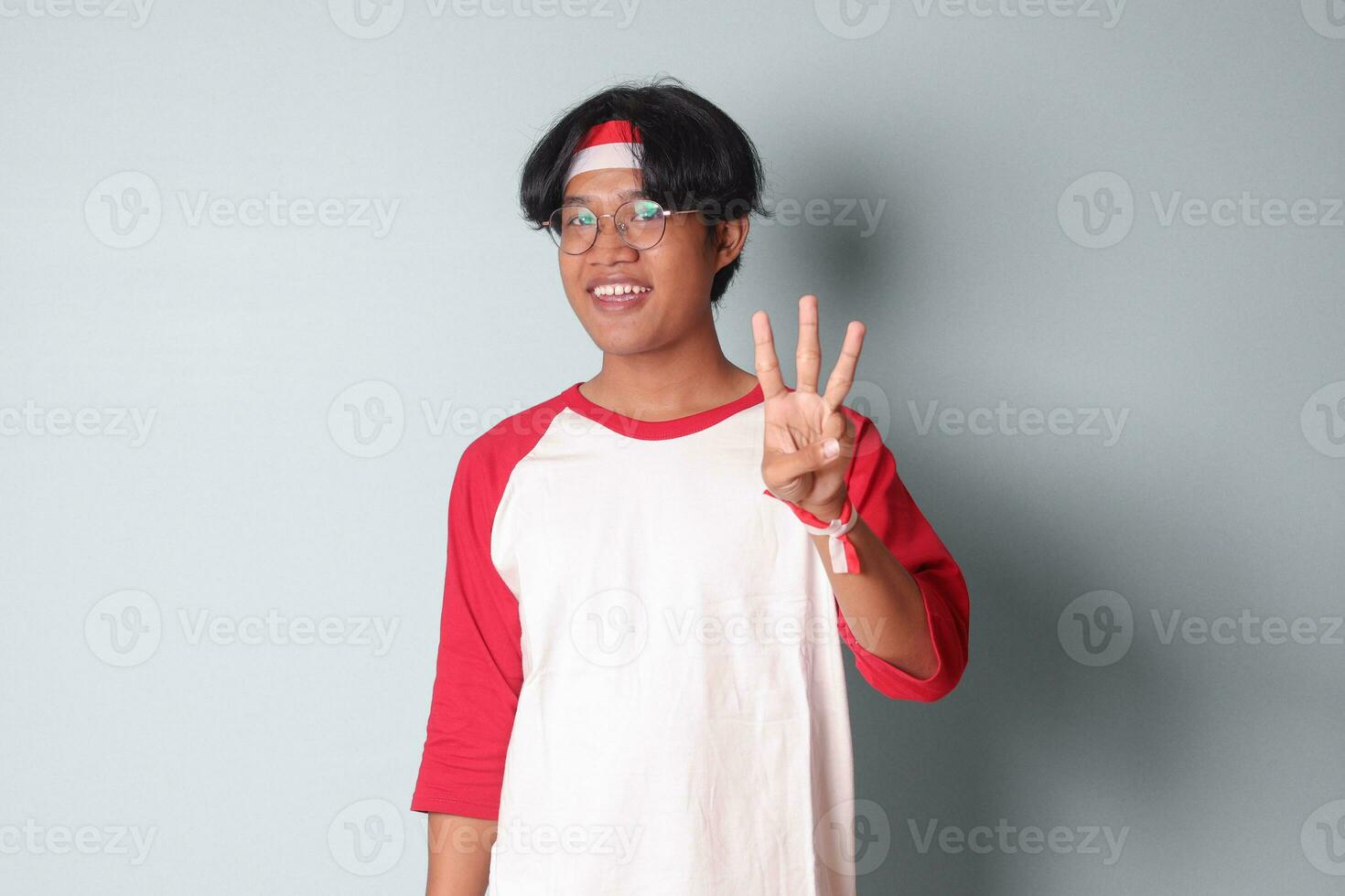 Portrait of attractive Asian man in t-shirt with red and white ribbon on head, counting three with fingers. Isolated image on gray background photo