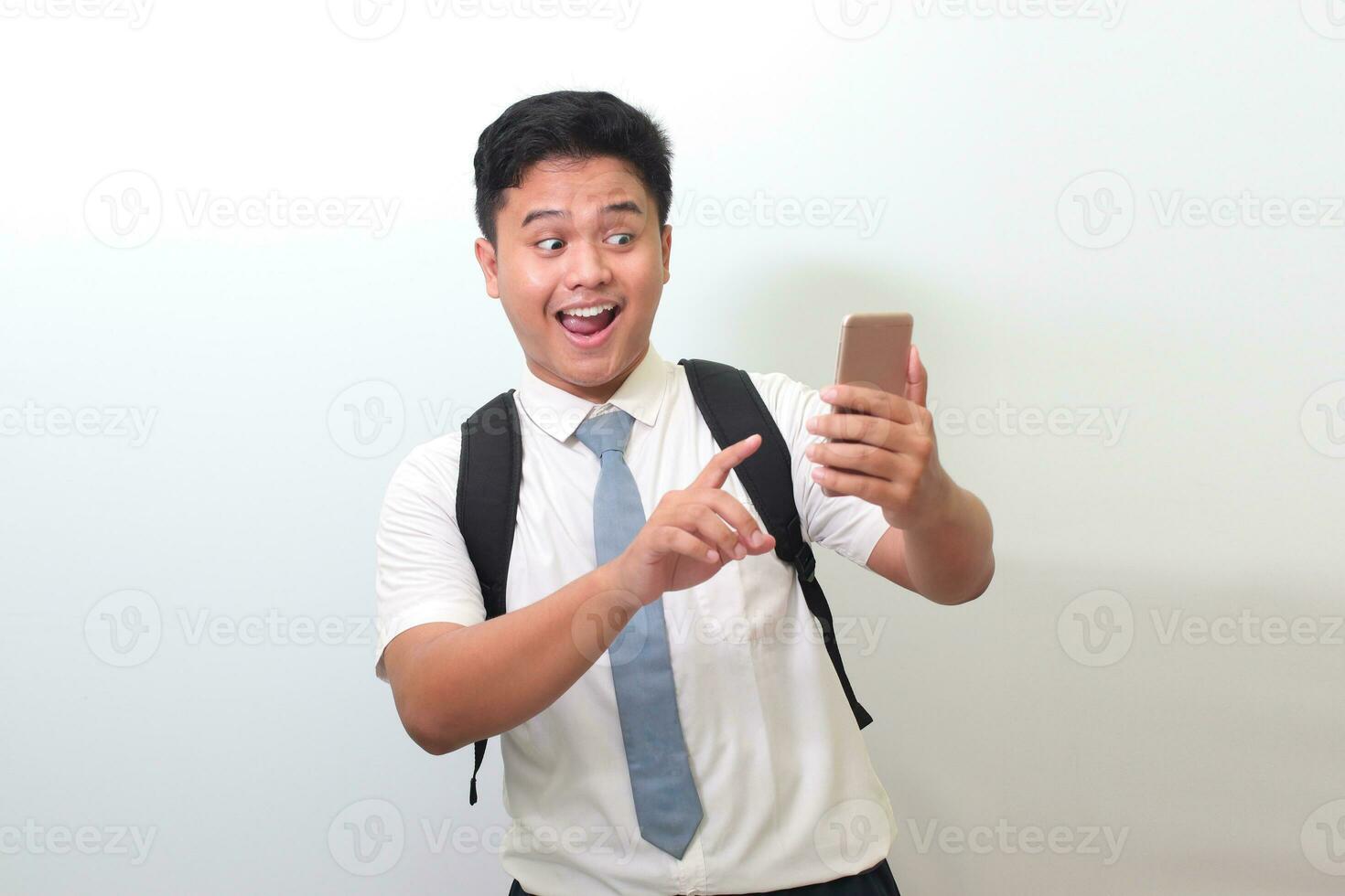 Indonesian senior high school student wearing white shirt uniform with gray tie showing shocked face expression while holding a mobile phone. Isolated image on white background photo