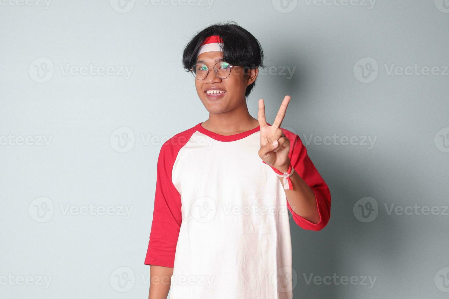 Portrait of attractive Asian man in t-shirt with red and white ribbon on head, counting two with fingers. Isolated image on gray background photo