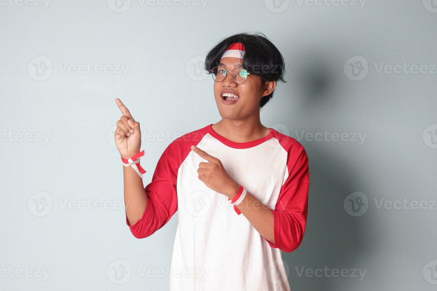 Portrait of attractive Asian man in t-shirt with red and white ribbon on head, pointing at something with finger. Isolated image on gray background photo