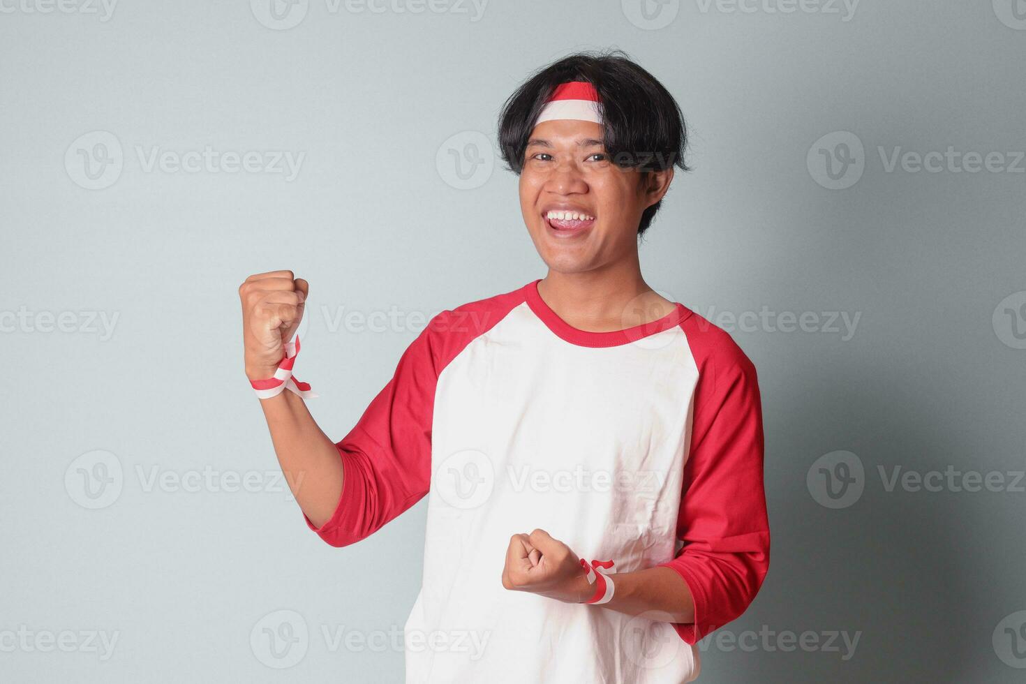 Portrait of attractive Asian man in t-shirt with red and white ribbon on head, raising his fist, celebrating Indonesia's independence day. Isolated image on gray background photo