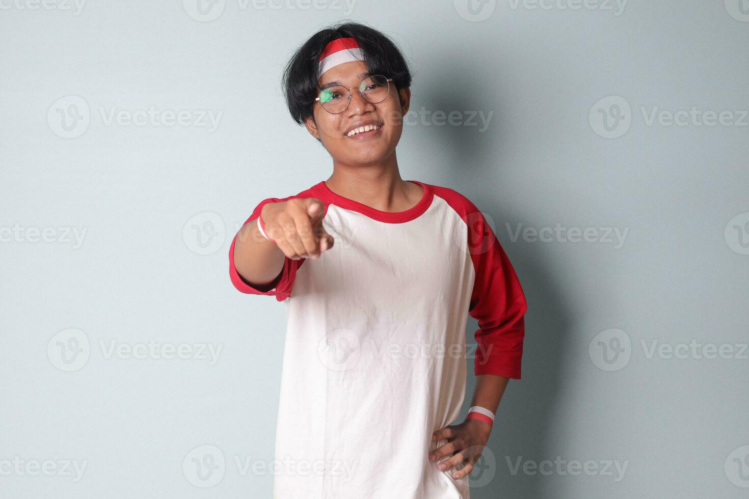 Portrait of attractive Asian man in t-shirt with red and white ribbon on head, pointing forward at the camera. Isolated image on gray background photo