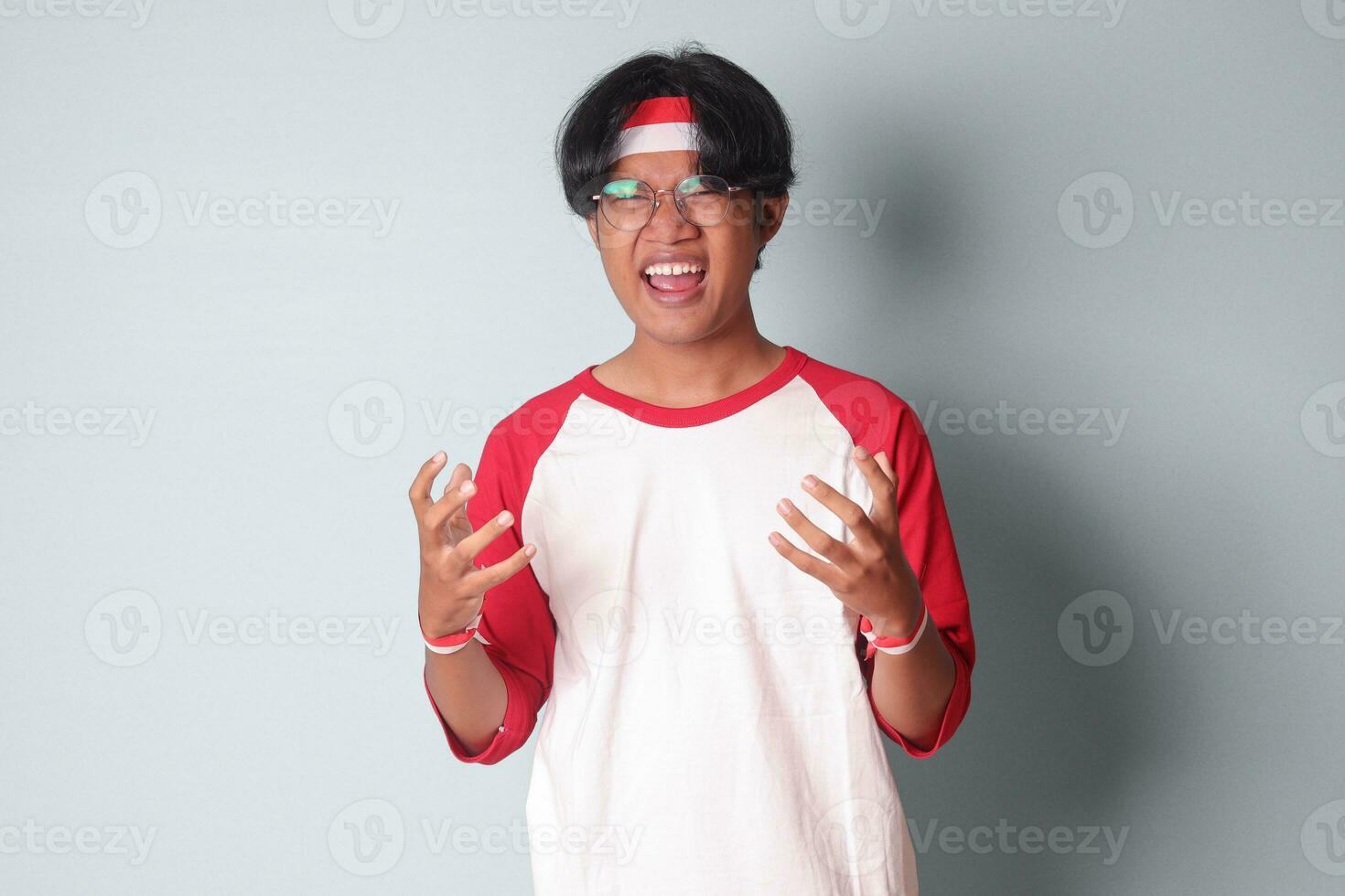 Portrait of attractive Asian man in t-shirt with red and white ribbon on head, shaking hands frustrated, feeling angry and mad at someone. Isolated image on gray background photo